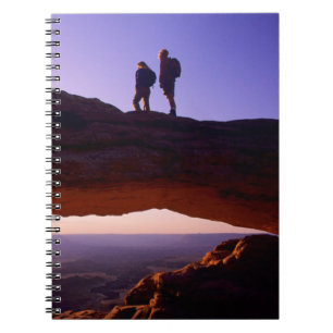 A couple watches sunrise from atop Mesa Arch in Notebook