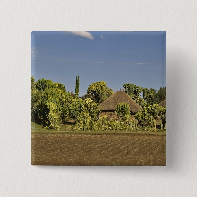 A farmed field in front of thatched roof houses 2 inch square button (Front)