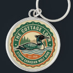 Cottage Loon Stupid Canadian Wolf Bird  Keychain<br><div class="desc">Circular badge with a central illustration of a waterfowl floating on stylized blue and cream waves. The bird features a dark head, a pointed beak, and a distinct white checkered pattern on its back. The background consists of green pine tree silhouettes against a pale sky. The design is framed by...</div>