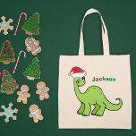 Cute Christmas Dinosaur Custom Green Kids Name Tote Bag<br><div class="desc">Awesome green dinosaur looking very cute in a holiday red Santa hat with a little smile. Adorable custom kids tote bag gift for your little boy or girl.</div>