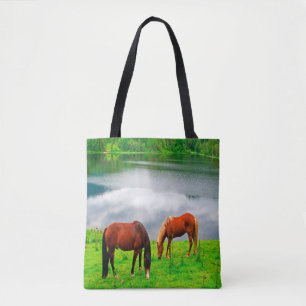 Horses grazing meadow with a forest lake tote bag