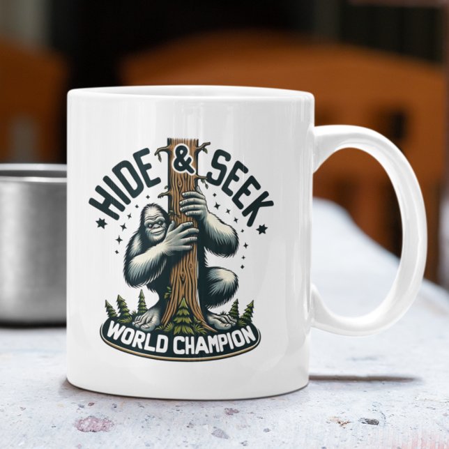 Mug Funny Cacher & Rechercher Champion du Monde Bigfoo (White mug with Bigfoot "Hide & Seek World Champion" design on an outdoor table.)
