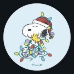 Peanuts | Christmas Love and Lights Classic Round Sticker<br><div class="desc">This circular Peanuts sticker features a pale blue background. It shows Snoopy, the famous beagle, sitting down and happily tangled in a string of colourful Christmas lights. He is wearing a festive, striped Santa hat. Nestled securely in his embrace is his best friend, Woodstock. This officially licensed sticker is a...</div>