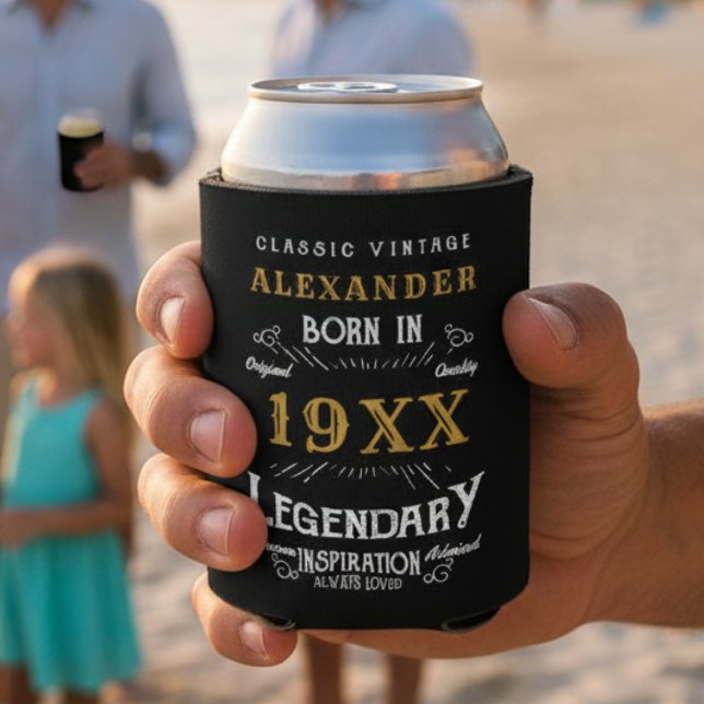 Personalized Birthday Add Your Name Year Legendary Can Cooler (Black and gold can cooler in a hand with a beach background.)