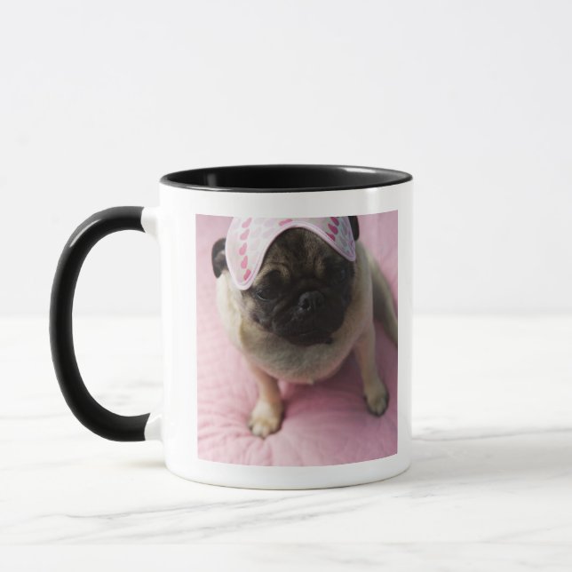 Pug dog with eye mask on head sitting on bed, mug (Left)