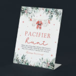 Red Christmas Santa Baby Pacifier Hunt Baby Shower Pedestal Sign<br><div class="desc">The Red Christmas Santa Baby Pacifier Hunt Baby Shower Game Sign adds a touch of holiday fun and excitement to your baby shower. Featuring a white background with festive red snowflakes, cheerful red berries, winter eucalyptus greenery, and Santa-themed baby clothes, this sign invites guests to join the "Pacifier Hunt" game....</div>