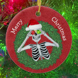 Skeleton in Pine Christmas Wreath Red Hat Funny Ceramic Ornament<br><div class="desc">This hanging ceramic ornament has a funny skeleton holding ornaments with snowflakes up to his eyes wearing a red Santa hat inside a green pin wreath on a deep red background.  Customize your text on the back with the and the Merry Christmas on front with the templates.</div>