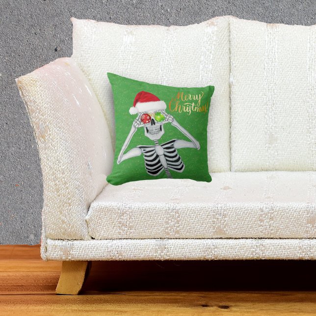 Smiling Skeleton Holding Ornaments to Eyes Green Throw Pillow (Happy Merry Christmas Skeleton in Santa hat with ornaments on green throw pillow.)