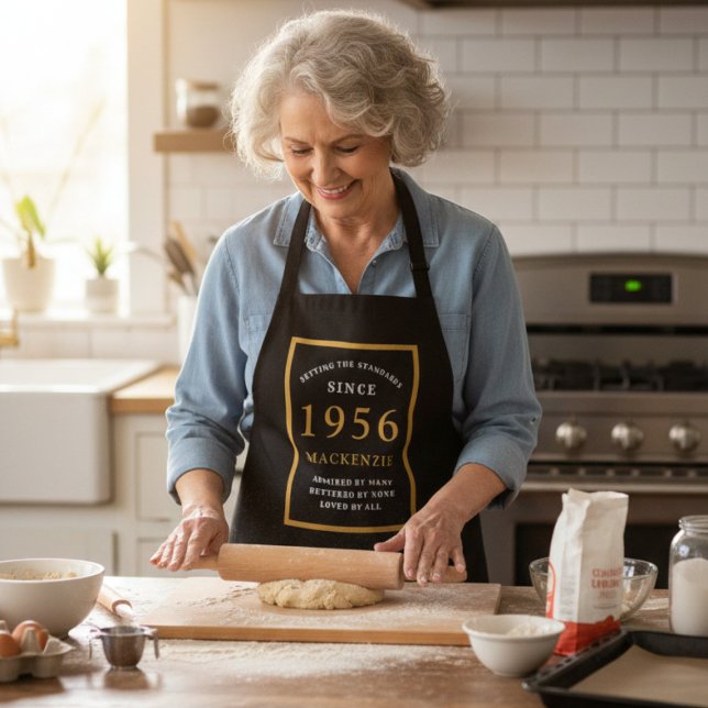 Tablier 70e anniversaire 1954 Nom Elegant Black Gold Chic (1956 apron worn by a woman baking in a kitchen.)