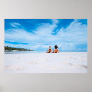 Two women lying on white sand facing beach under b poster