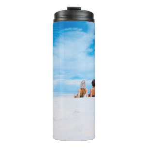 Two women lying on white sand facing beach under b thermal tumbler